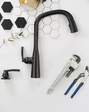 Single-handle Delta kitchen faucet and soap dispenser in matte black lie on white surface with installation tools and hexagon tile samples. 